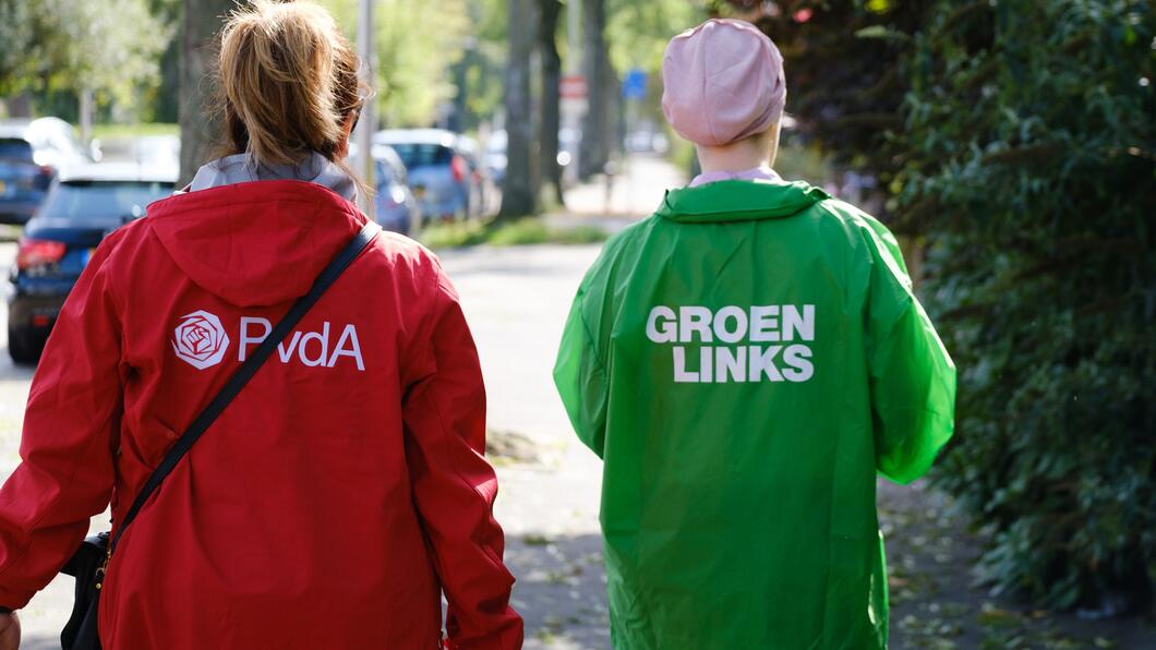 Twee campagnevoerders lopen naast elkaar. Eén van hen draagt een rode PvdA-jas, de ander een groene GroenLinks-jas. 